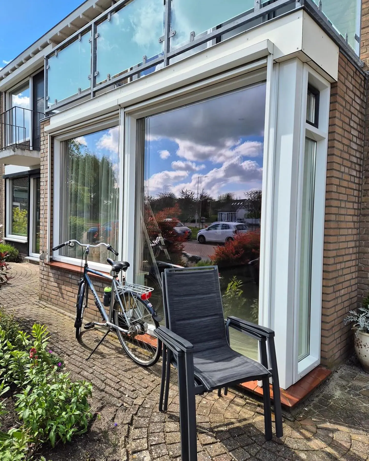 Witte kozijnen met grote ruiten en een smal zijraam op de hoek van een uitbouw, onder een balkon met glazen hekwerk.